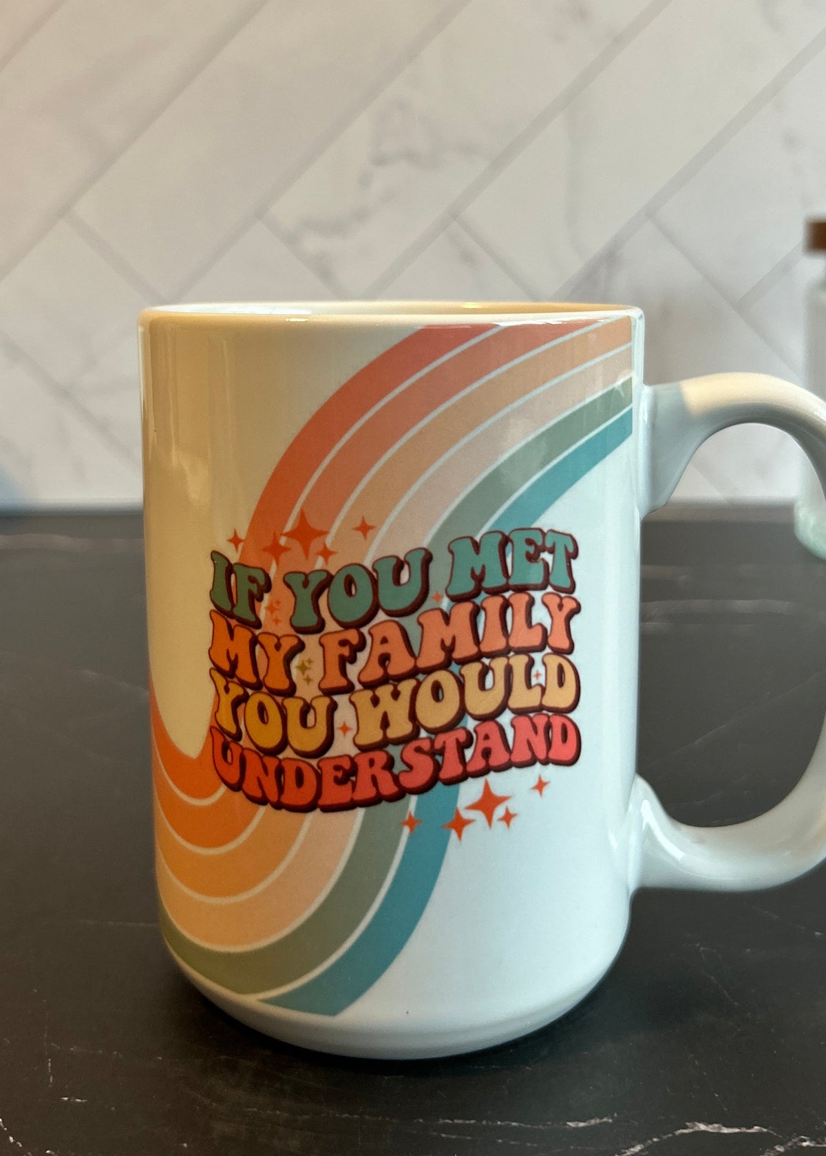 A white ceramic coffee mug with a colorful retro rainbow design and the text 'IF YOU MET MY FAMILY YOU WOULD UNDERSTAND' sitting on a dark countertop.