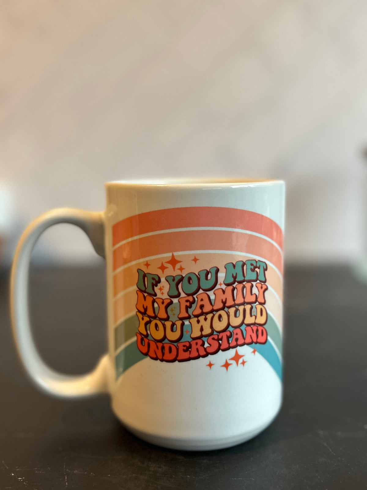 A white ceramic mug with a retro rainbow design and the text 'IF YOU MET MY FAMILY YOU WOULD UNDERSTAND' sitting on a dark countertop.