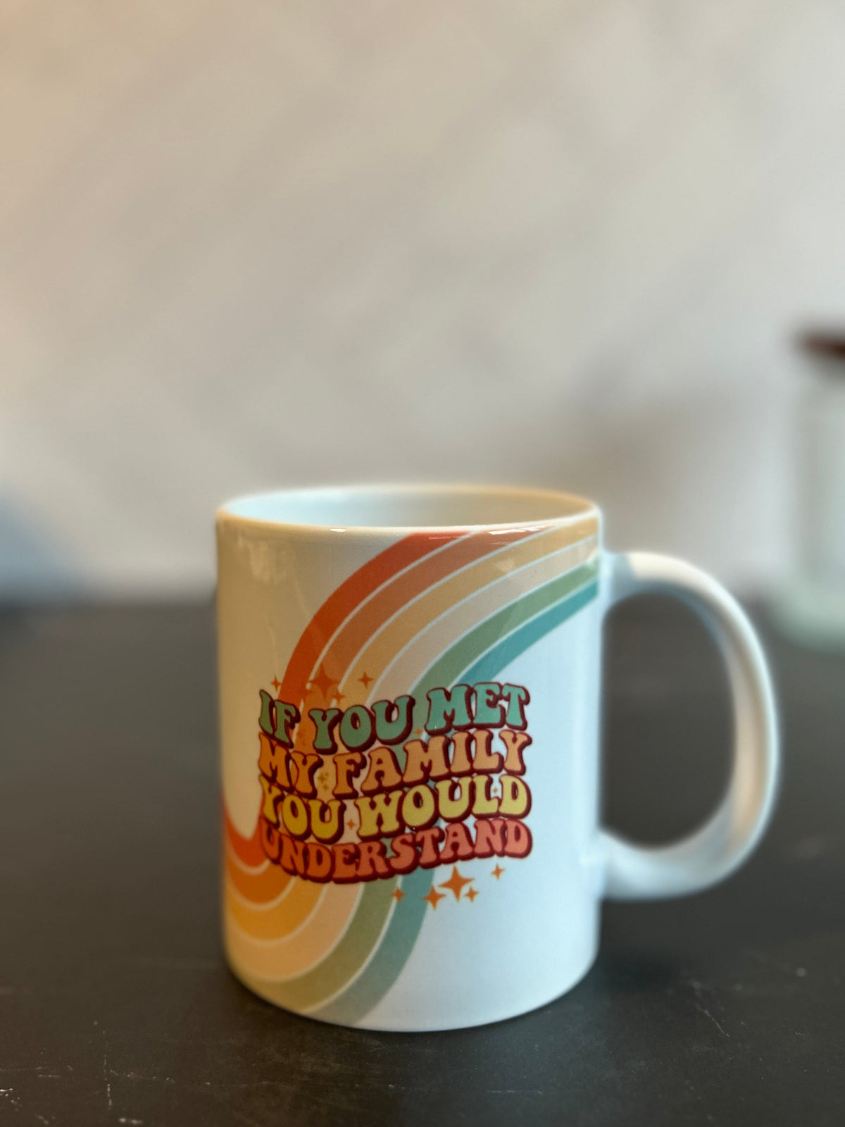 A white ceramic coffee mug with a retro rainbow design and the text 'IF YOU MET MY FAMILY YOU WOULD UNDERSTAND' sitting on a dark marble countertop.
