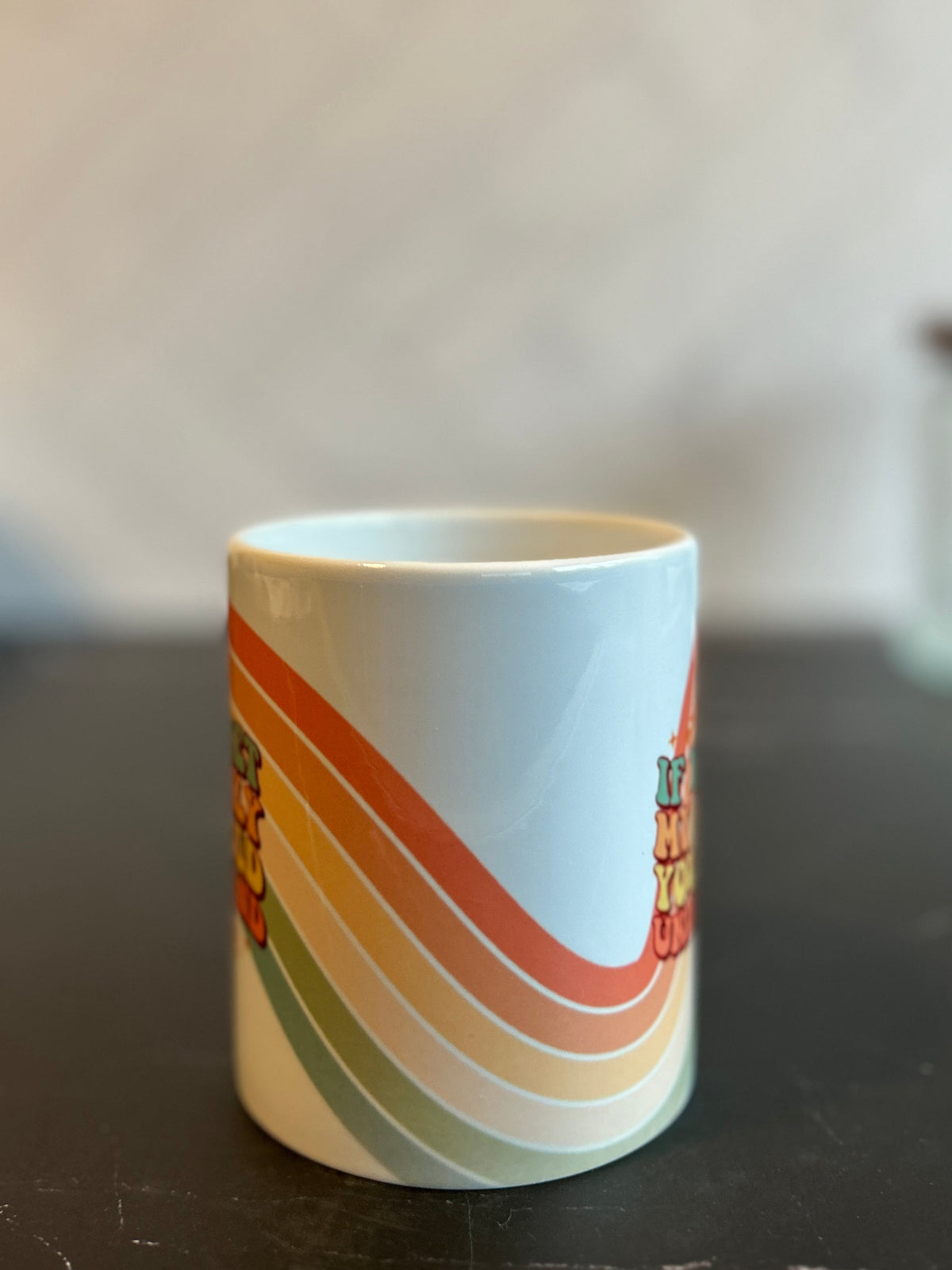 A white ceramic mug with a retro 1970s-style rainbow stripe pattern in orange, yellow, green, and red, sitting on a dark marble tabletop with a blurred background.