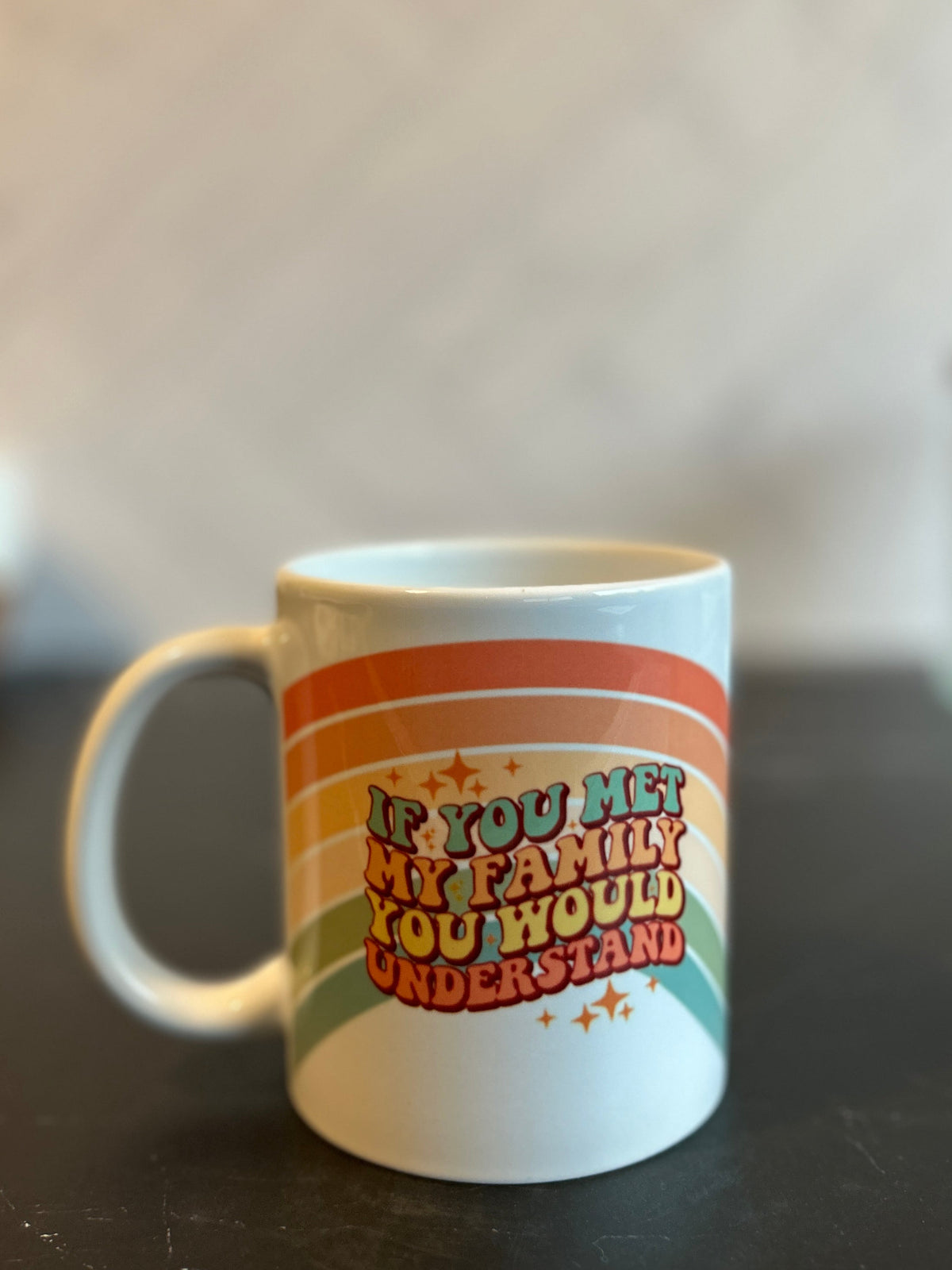 A white ceramic coffee mug with a retro rainbow design and the text 'IF YOU HATED MY FAMILY YOU WOULD UNDERSTAND' sitting on a dark countertop.