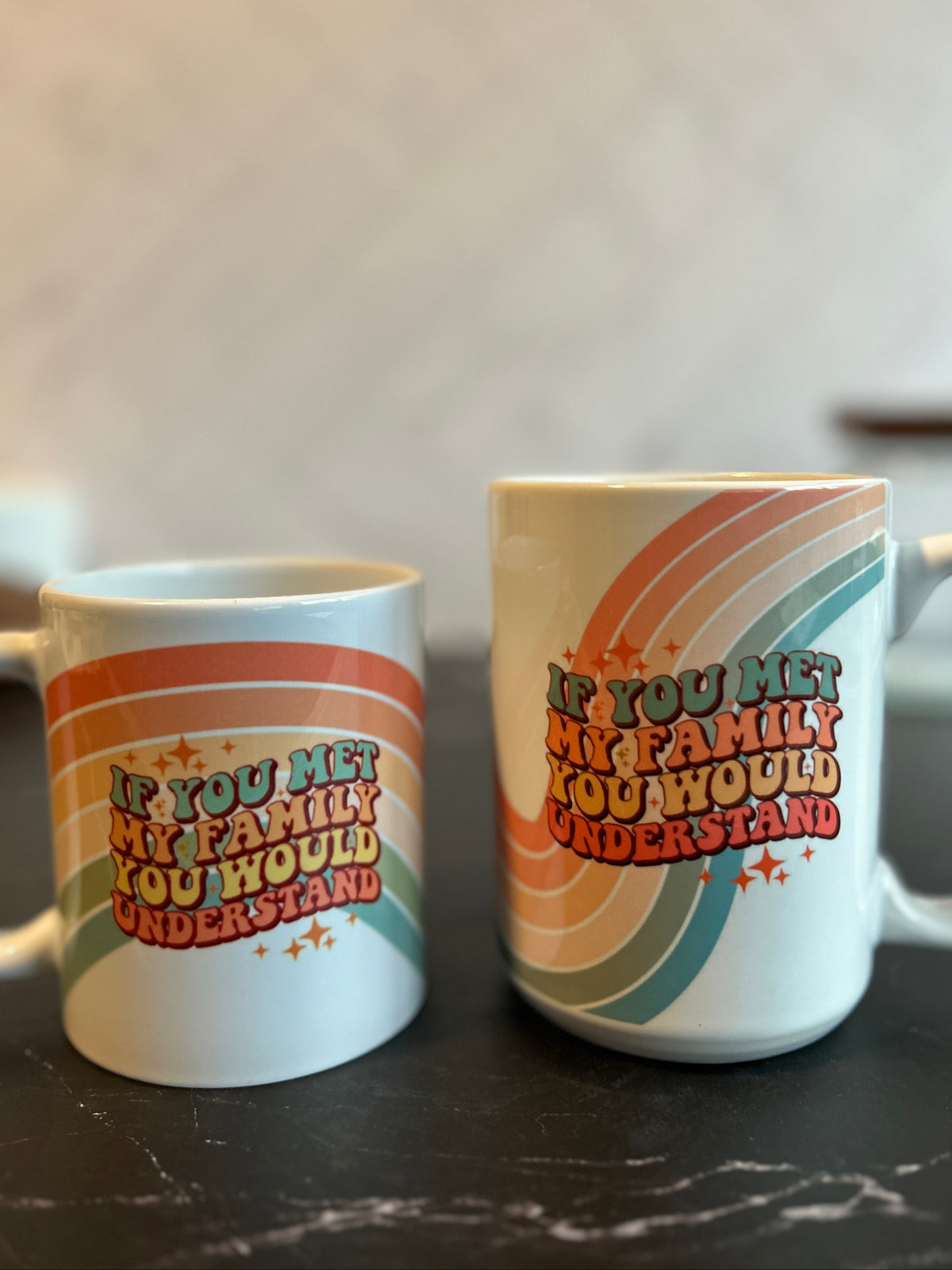 Two white ceramic mugs with retro rainbow stripes and the text 'IF YOU MET MY FAMILY YOU WOULD UNDERSTAND' on a dark marble surface against a blurred light background.