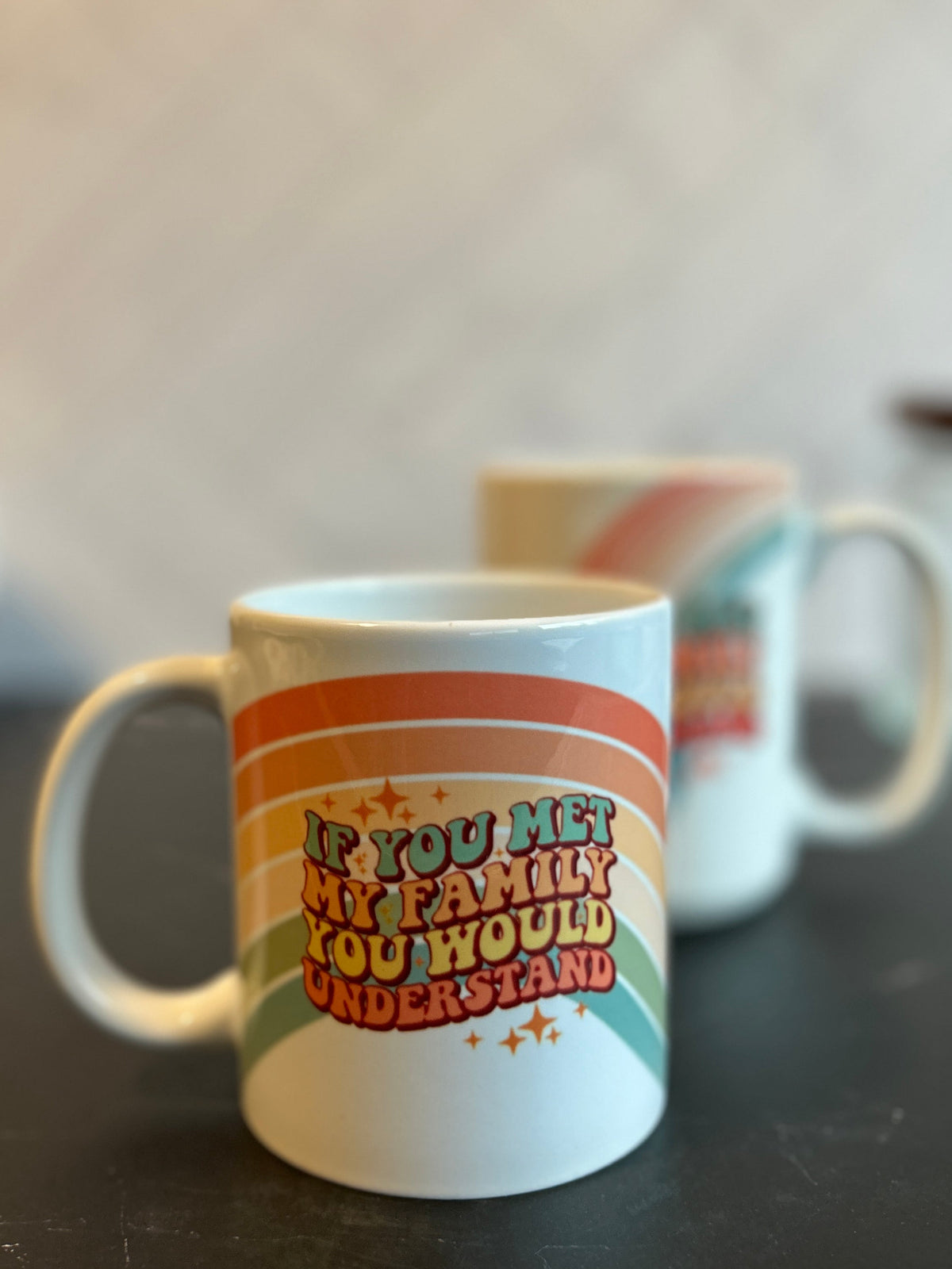 A white ceramic coffee mug with a retro rainbow design and the text 'IF YOU MET MY FAMILY YOU WOULD UNDERSTAND' sits on a dark countertop, with another mug blurred in the background.