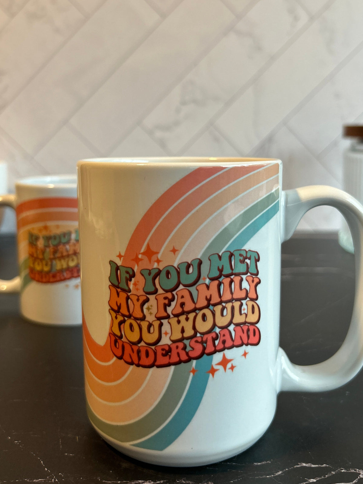 A white ceramic coffee mug with a colorful retro rainbow design and the text 'IF YOU MET MY FAMILY YOU WOULD UNDERSTAND' sitting on a dark countertop with a tiled backsplash behind it.