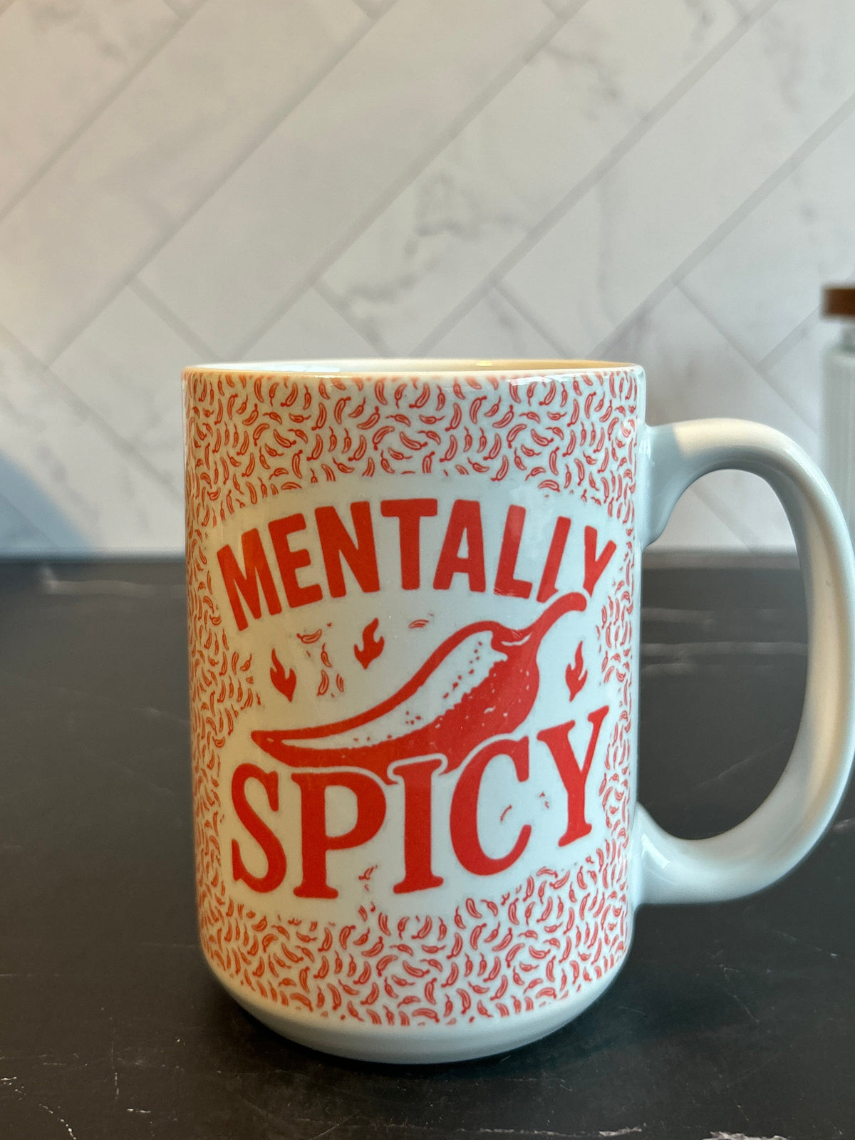 A white ceramic mug with a red patterned design and the words 'MENTALLY SPICY' printed on the front, sitting on a dark countertop with a tiled kitchen background.