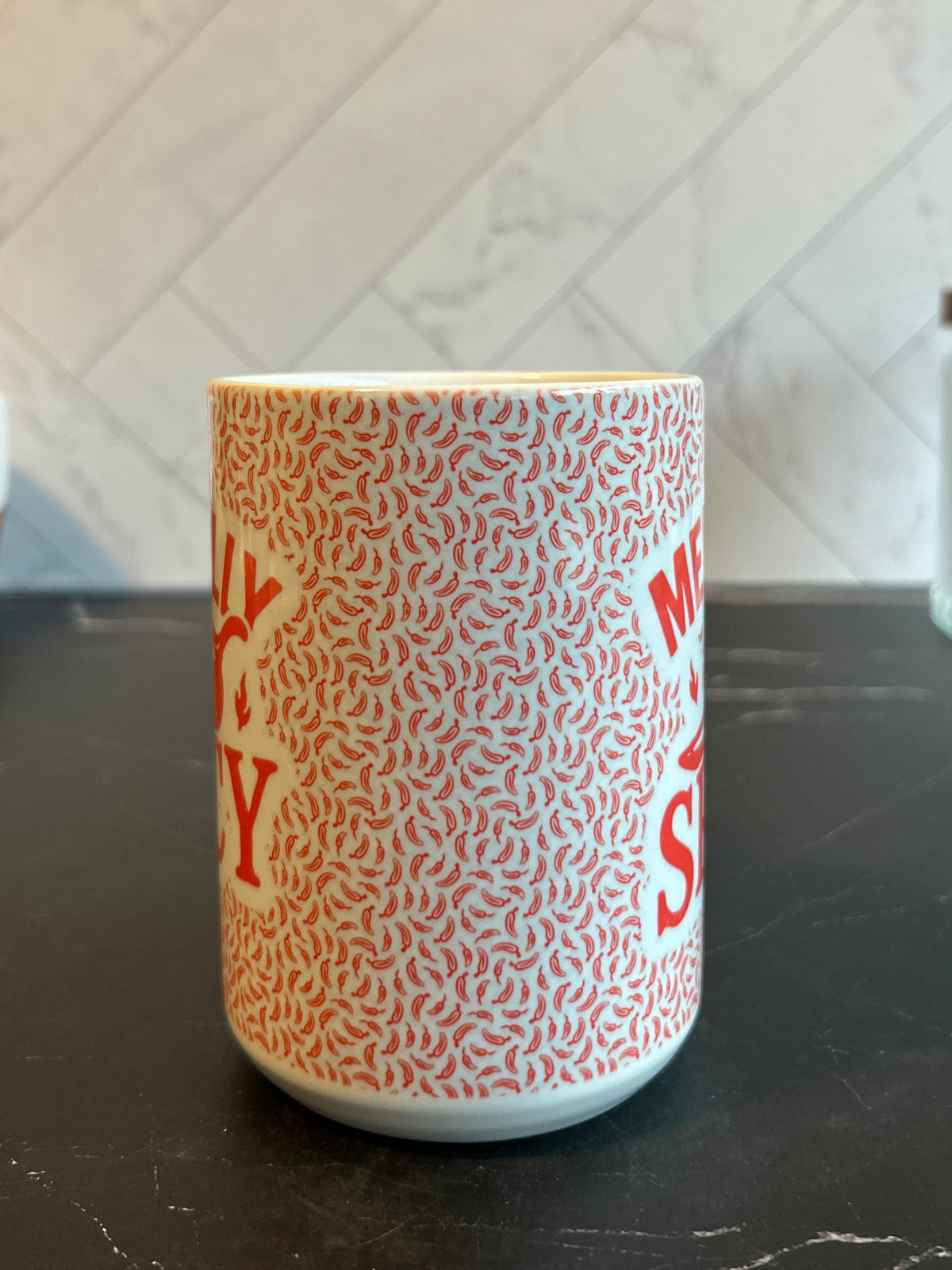 A white ceramic mug with a dense orange scribble pattern and red text reading 'WEDNESDAY' on the side, set on a dark countertop with a tiled wall background.