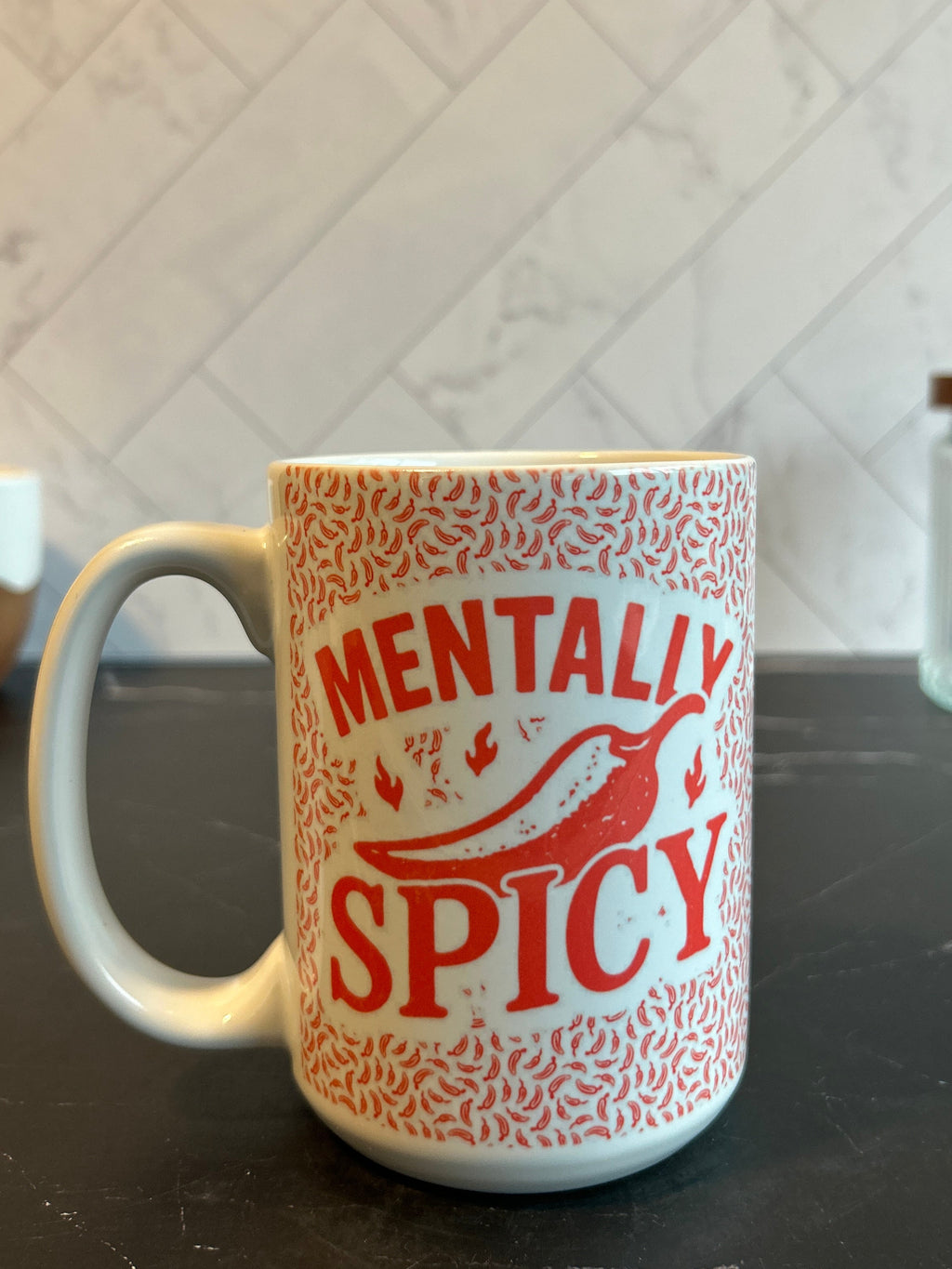 A white ceramic mug with a red patterned design and the words 'MENTALLY SPICY' printed on the front, sitting on a dark countertop with a tiled kitchen background.