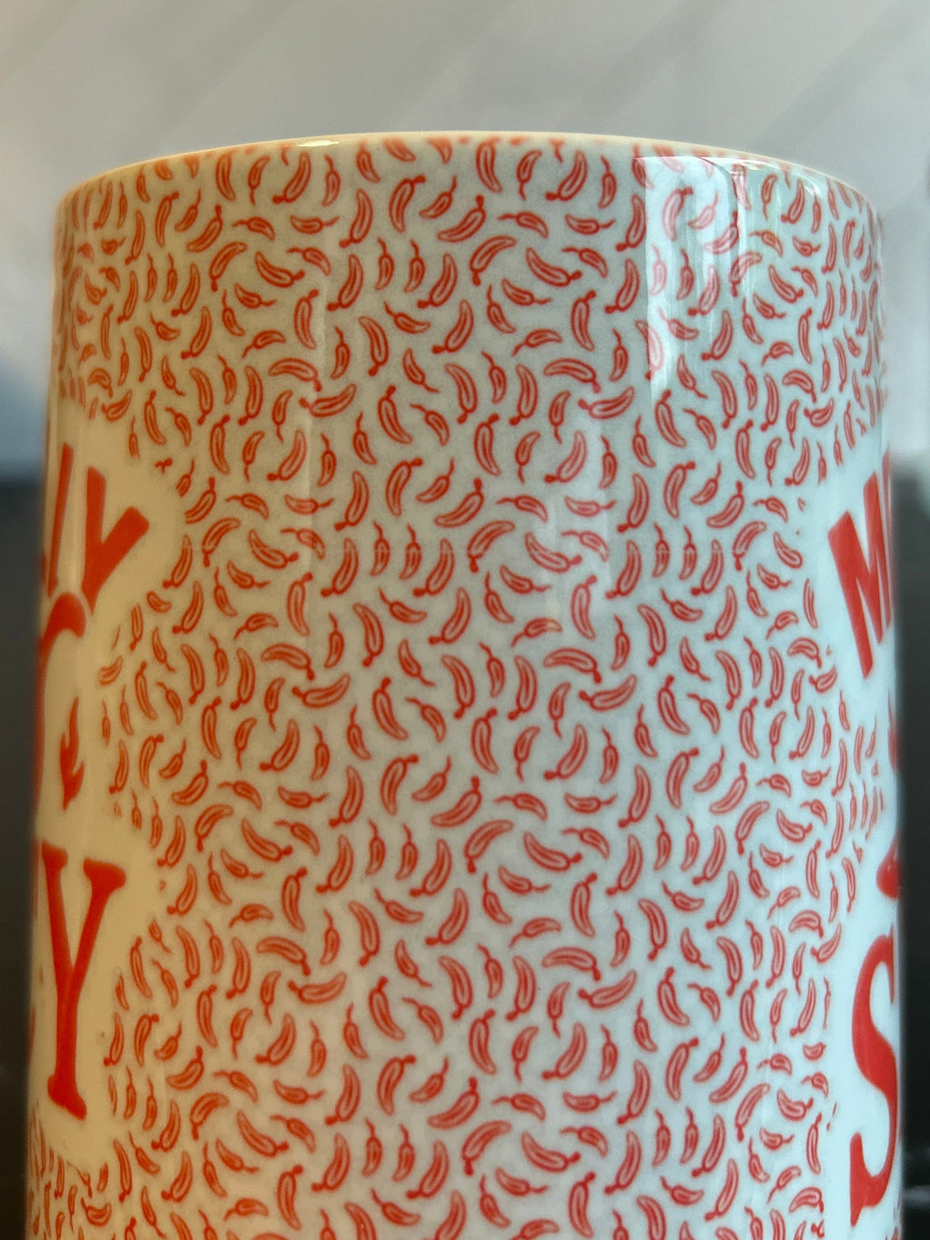 A close-up of a white ceramic mug with a repeating red chili pepper pattern and partial red lettering, set on a dark countertop with a blurred kitchen background.