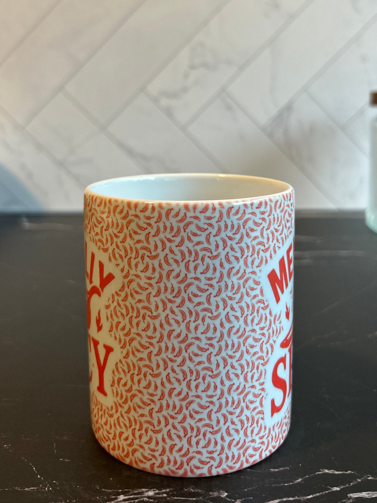 A white ceramic mug with a dense red abstract pattern and partial red text, standing on a dark countertop with a tiled backsplash behind it.