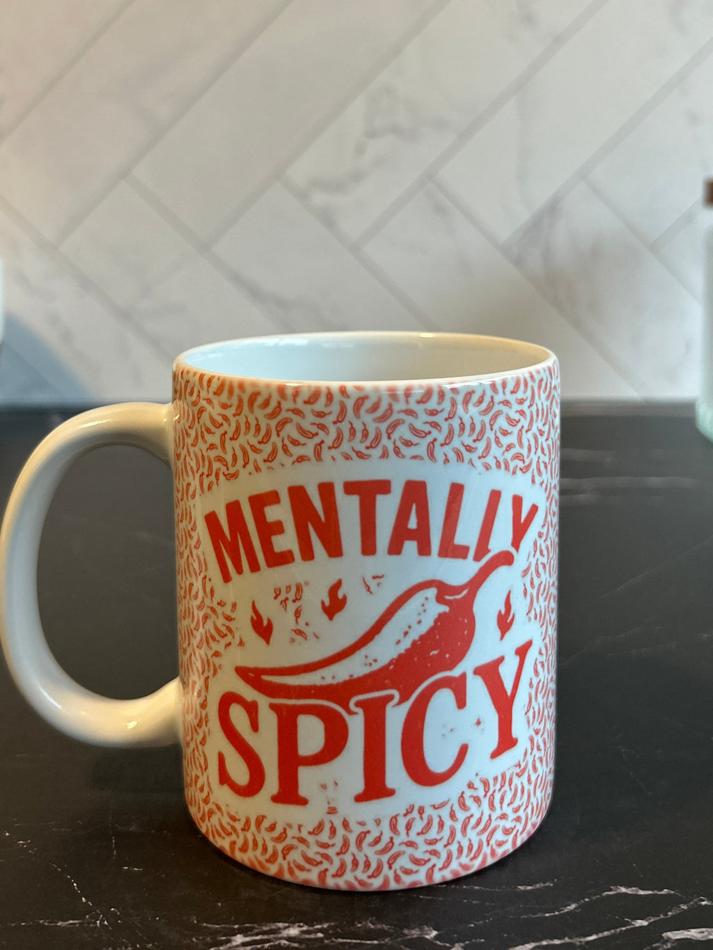 A ceramic mug with a white handle and base, decorated with a red and orange pattern and the words 'MENTALLY SPICY' in red, sitting on a dark countertop with a tiled backsplash behind it.