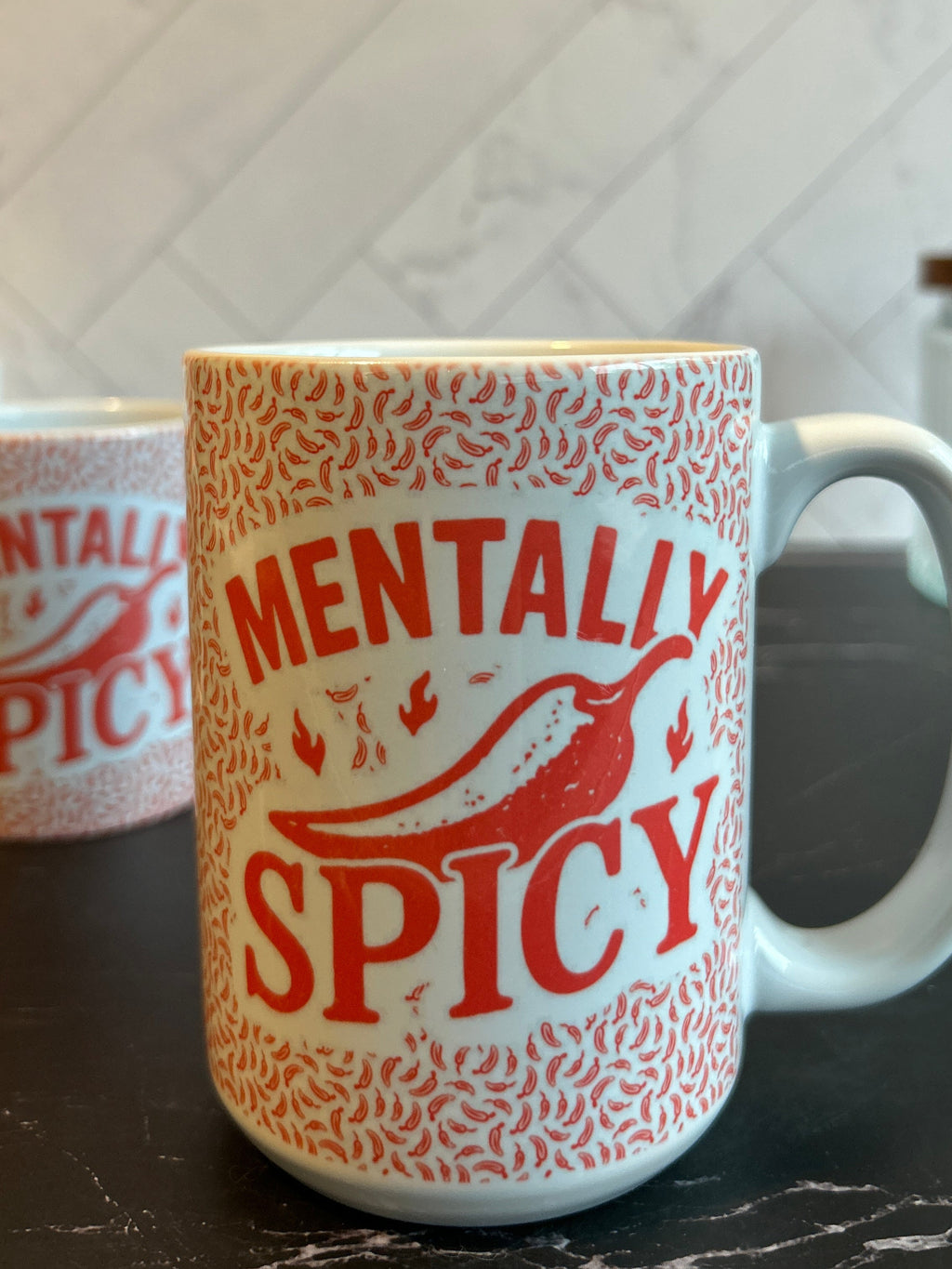 A white ceramic mug with a red patterned border and the words 'MENTALLY SPICY' printed in red, featuring a red chili pepper graphic. A second mug is partially visible in the background on a dark countertop against a light tiled wall.