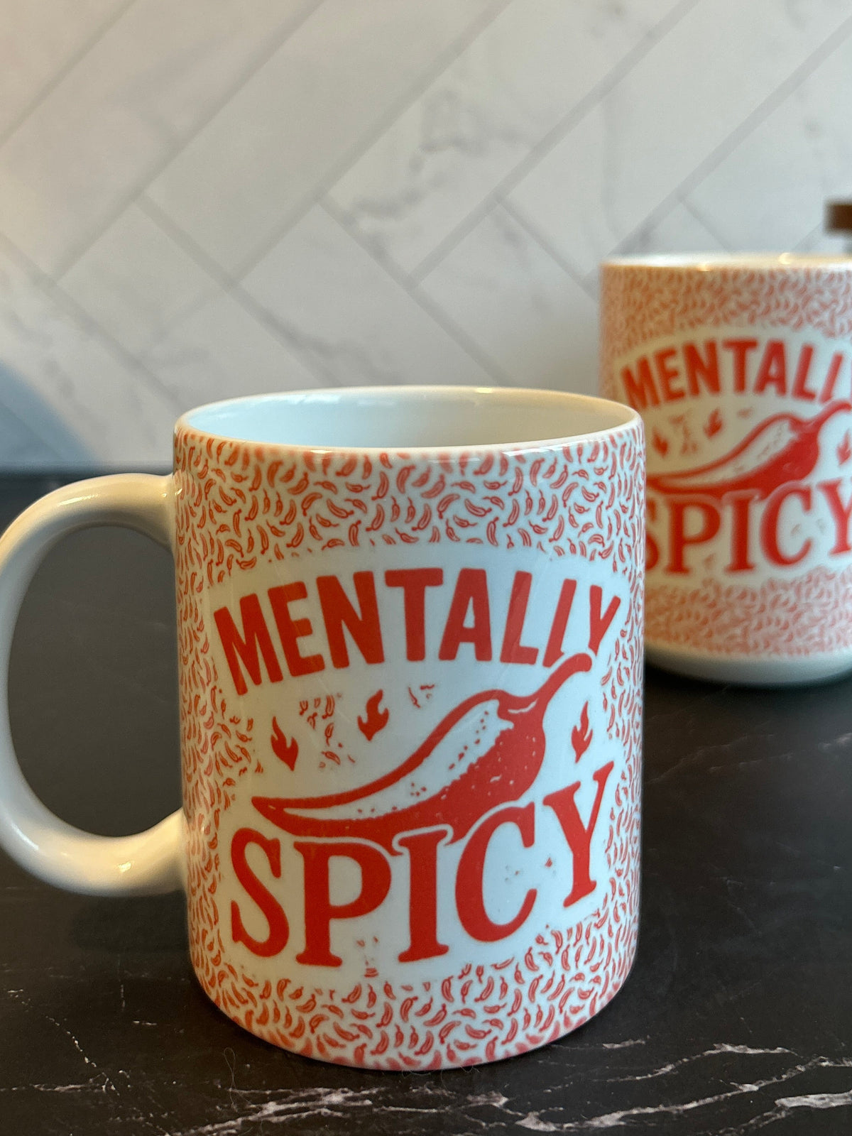 A white ceramic mug with a red and orange patterned design and the words 'MENTALLY SPICY' printed on the front, sitting on a dark countertop with a tiled wall in the background.