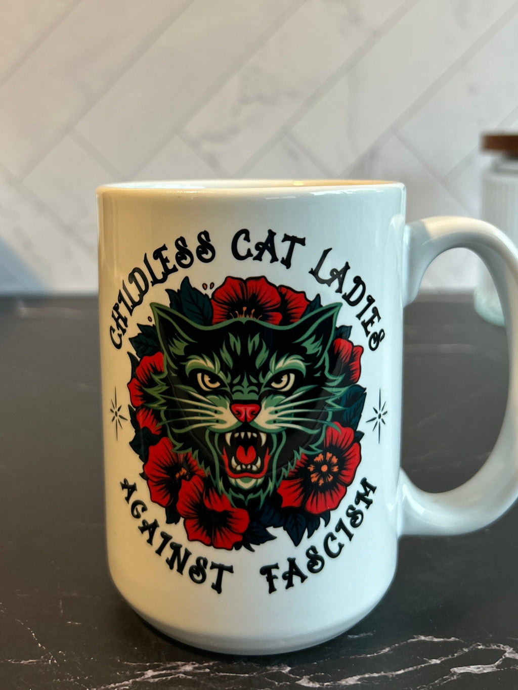 A white ceramic mug with a bold graphic of a green cat surrounded by red flowers and the text 'GODLESS CAT LADIES AGAINST FASCISM', set on a dark countertop with a tiled kitchen background.