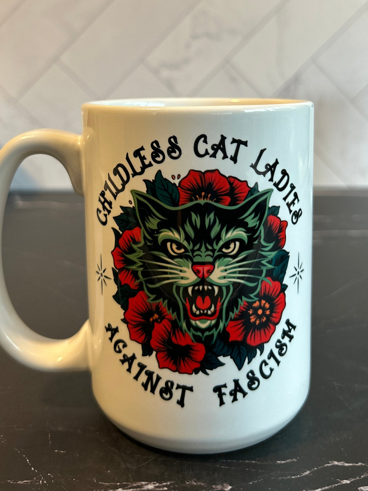 A white ceramic mug with a graphic of a snarling cat surrounded by red roses and the text 'COTILESS CAT LADIES AGAINST FASCISM', set on a dark countertop with a tiled kitchen background.