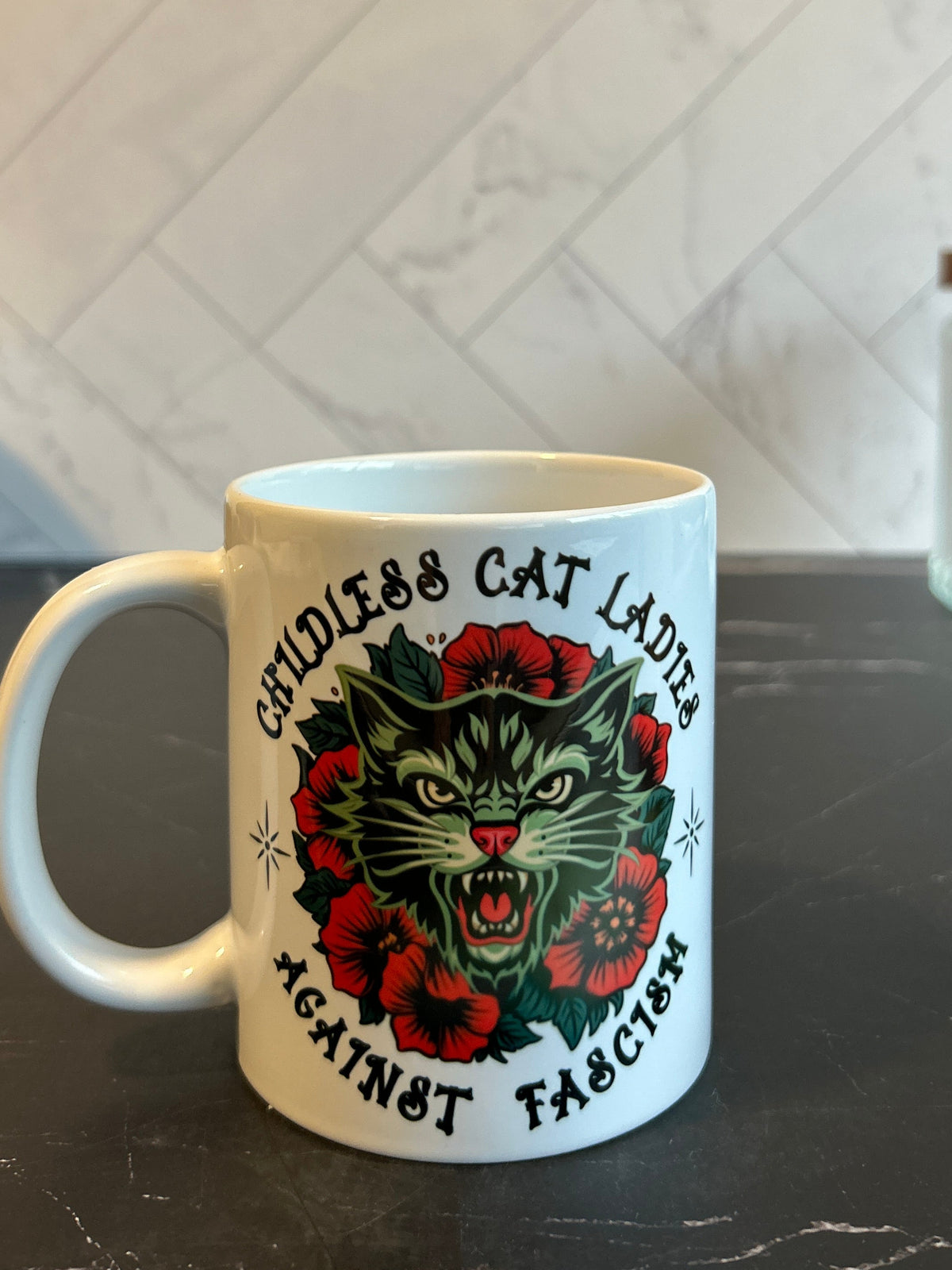 A white ceramic coffee mug with a graphic of a green cat surrounded by red roses and the text 'Cruelty Free Cat Lovers Against Fiction', set on a dark countertop with a tiled backsplash.