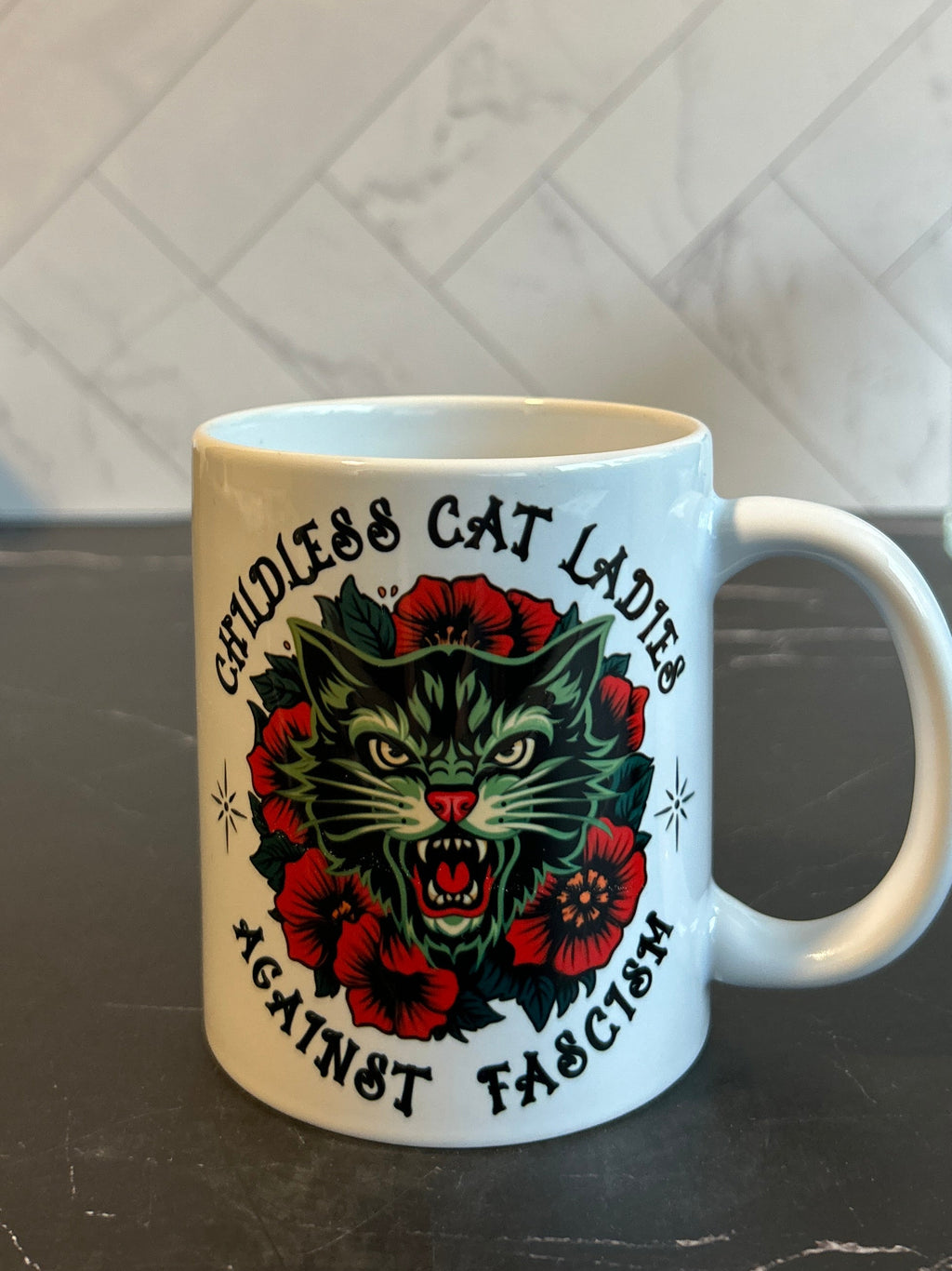 A white ceramic coffee mug with a graphic of a green cat surrounded by red roses and the text 'Cruel & Cat Lady's Against Fascism', set on a dark marble countertop with a tiled backsplash in the background.