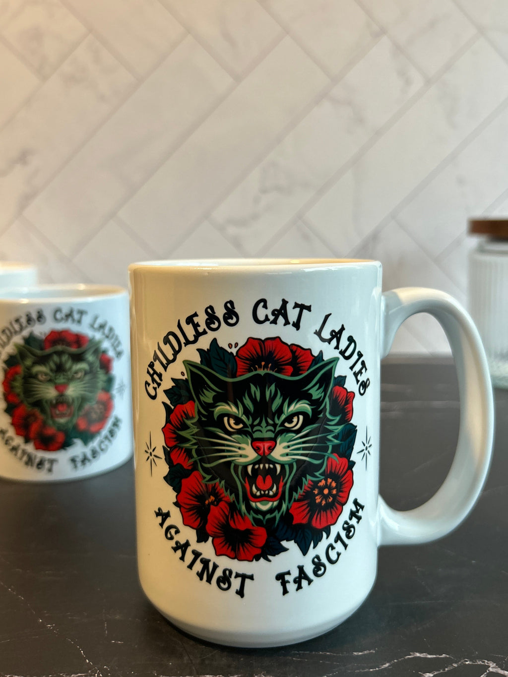 A white ceramic mug with a graphic of a roaring cat surrounded by red flowers and the text 'Cruelless Cat Lady' and 'Against Fascism', sitting on a dark countertop with a tiled wall in the background.