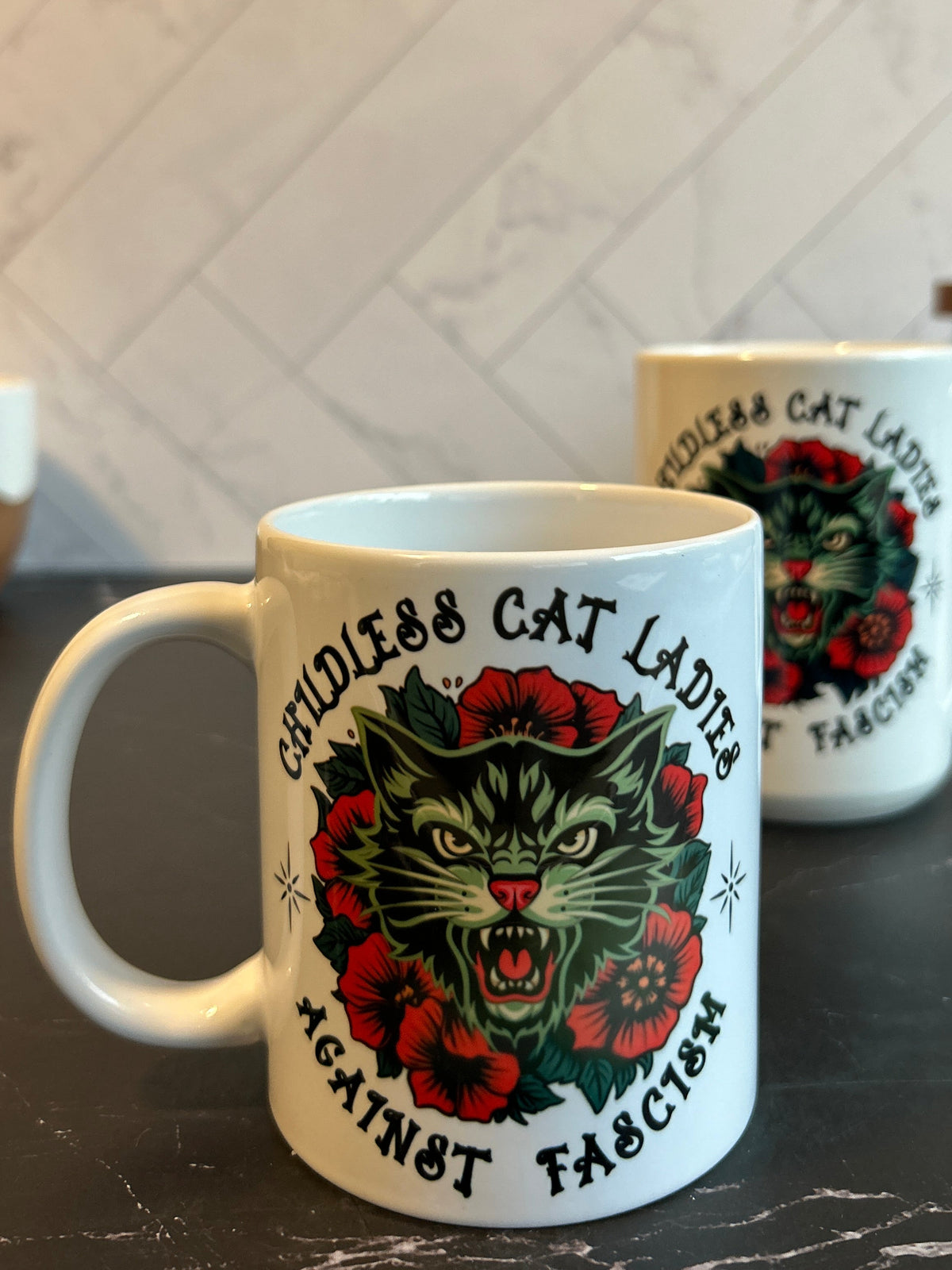 A white ceramic mug with a graphic of a roaring cat surrounded by red flowers and the text 'WEEDLESS CAT LADIES AGAINST FASCISM', shown on a dark countertop with a tiled backsplash in the background.
