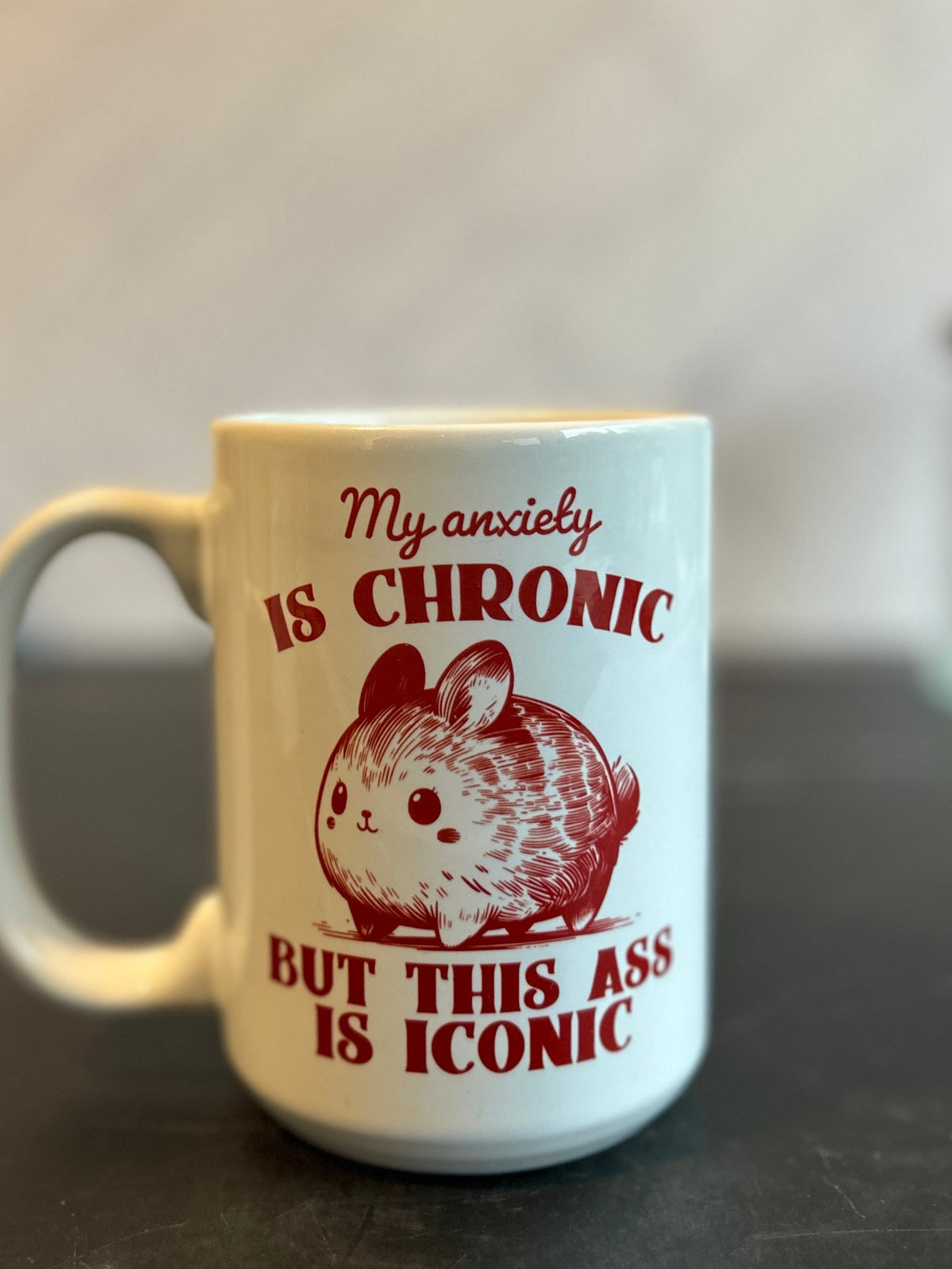 A white ceramic mug with a red graphic of a hamster and the text 'My anxiety IS CHRONIC BUT THIS ASS IS ICONIC', placed on a dark countertop with a blurred indoor background.