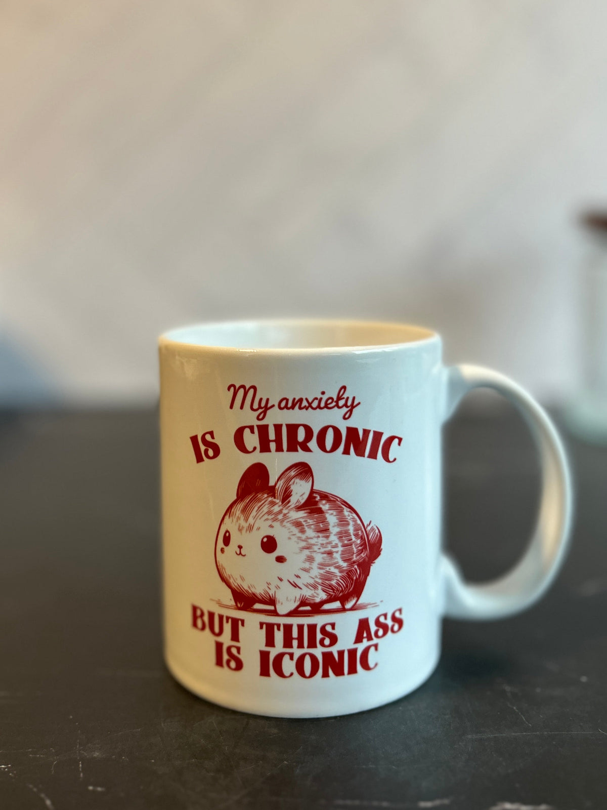 A white ceramic coffee mug with a humorous red graphic and text reading 'My anxiety IS CHRONIC BUT THIS ASS IS ICONIC' alongside a hamster illustration, sitting on a dark marble countertop.