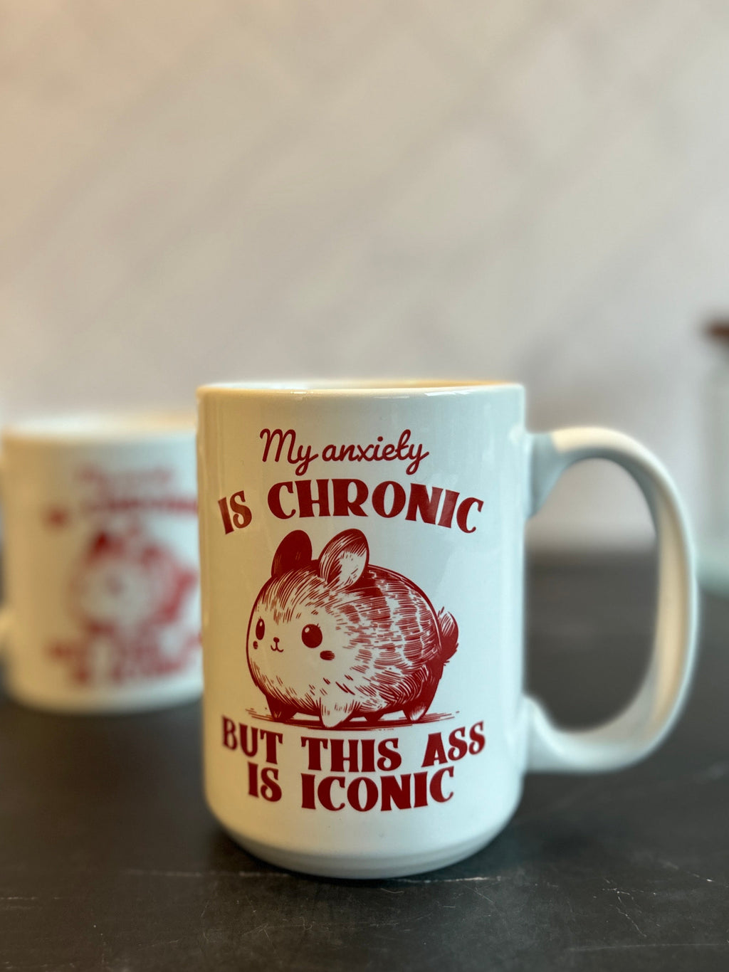 A white ceramic mug with a red graphic of a hamster and the text 'My anxiety IS CHRONIC BUT THIS ASS IS ICONIC' sitting on a dark countertop, with another blurred mug in the background.
