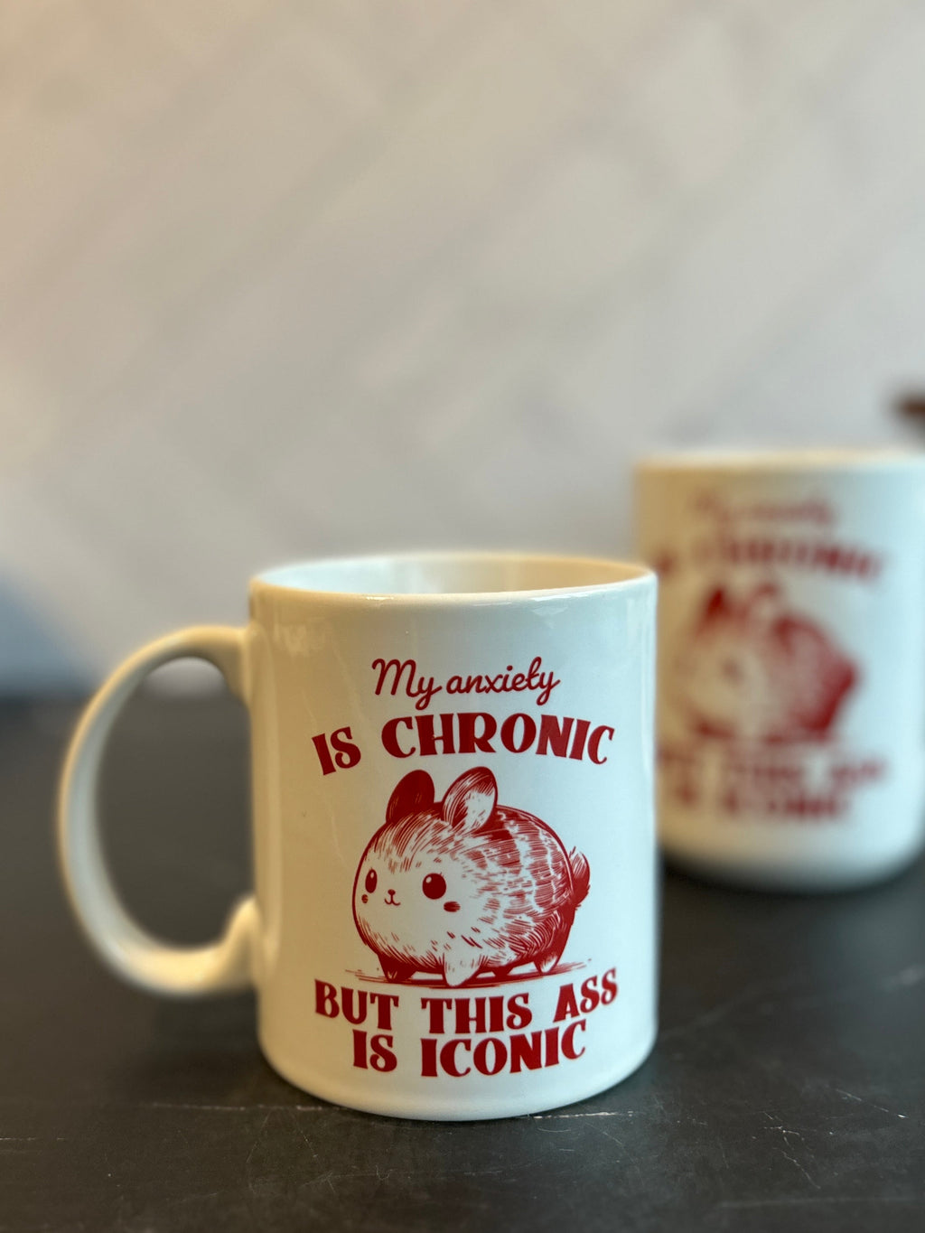 A white ceramic mug with red printed text and a hamster illustration sits on a dark marble countertop, with a second blurred mug in the background against a light wall.