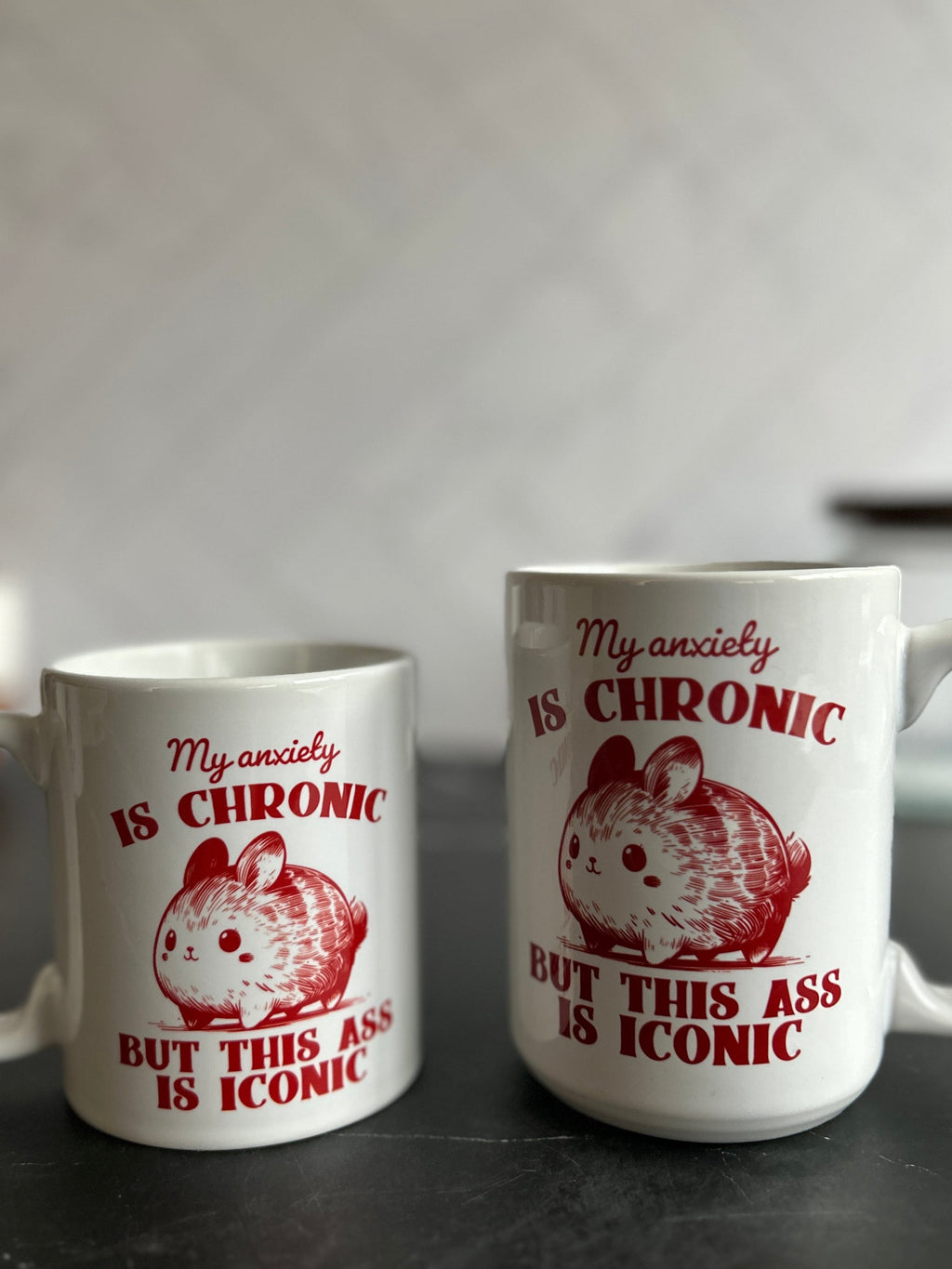 Two white ceramic mugs with red text and a hamster illustration, displayed on a dark marble countertop against a blurred gray background.