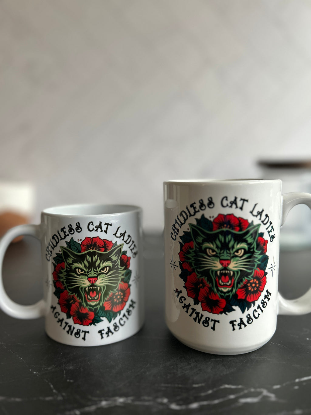 Two white ceramic mugs with a matching graphic print of a snarling green cat surrounded by red roses and the text 'Cruelty Free Cat Lads Against Fascism', displayed on a dark marble surface against a blurred light gray background.