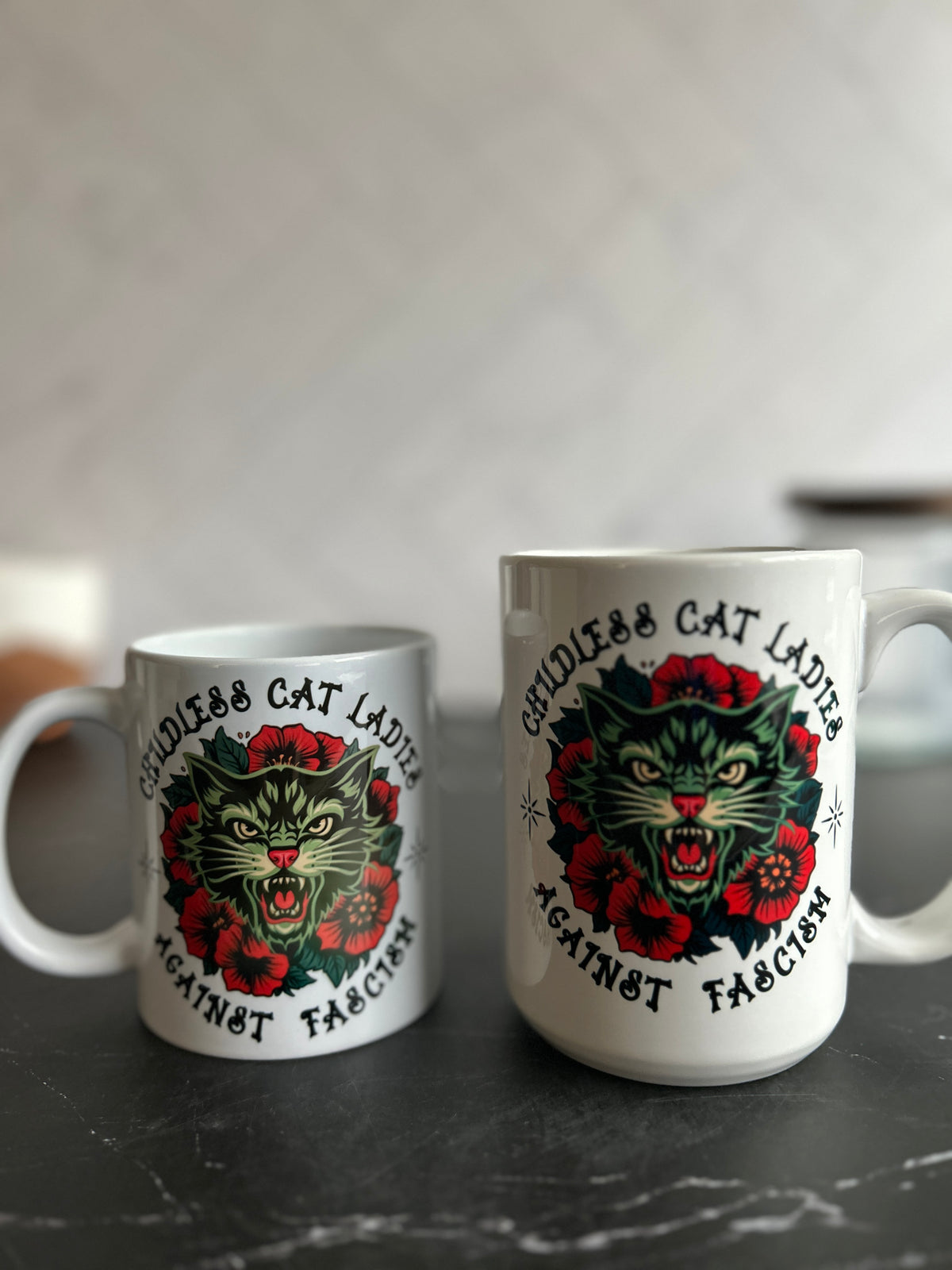 Two white ceramic mugs with a matching graphic print of a snarling green cat surrounded by red roses and the text 'Cruelty Free Cat Lads Against Fascism', displayed on a dark marble surface against a blurred light gray background.