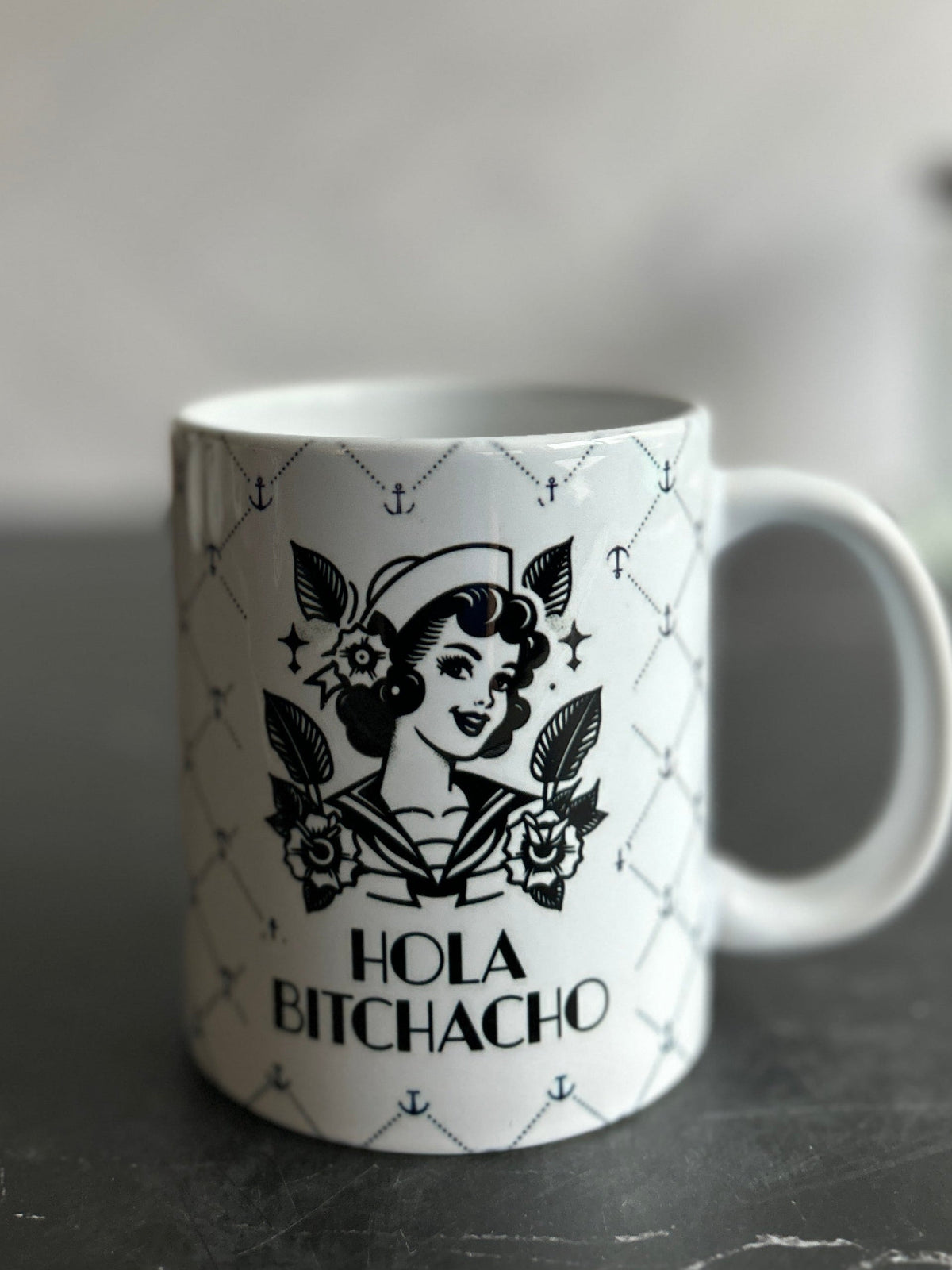A white ceramic mug with a black graphic of a retro pin-up woman and the text 'HOLA BITCHACHO' on a dark tabletop against a blurred gray background.