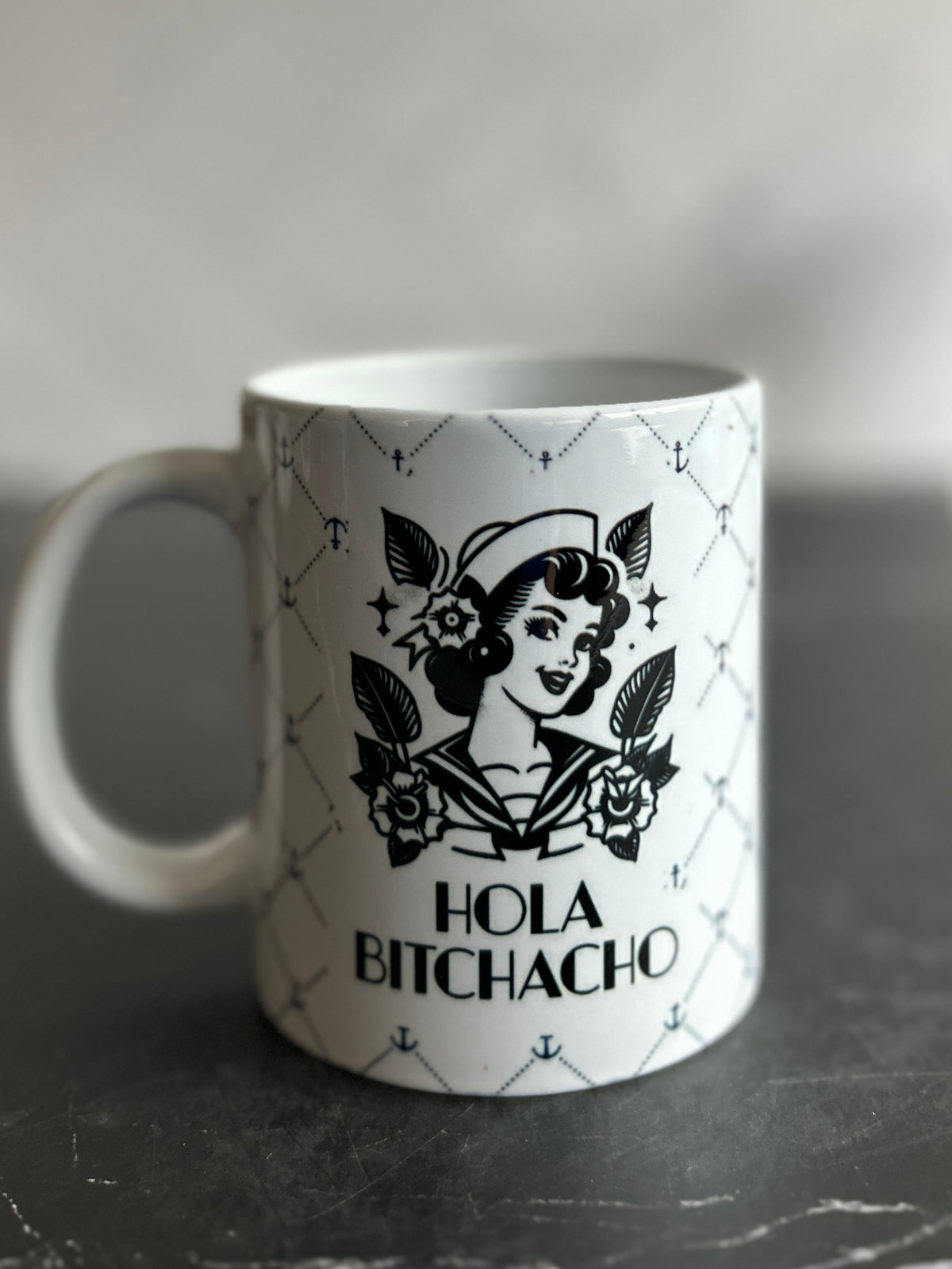 A white ceramic mug with a black graphic of a woman's profile, floral elements, and the text 'HOLA BITCHACHO' on a dark tabletop against a blurred light gray background.
